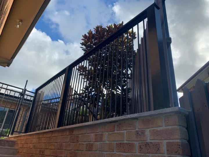 Black aluminium pool fence fixed along the top of a brick retaining wall, seen from below