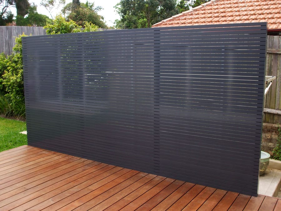 Freestanding charcoal aluminium slat privacy screen at the edge of a timber deck, in front of a neighbouring house