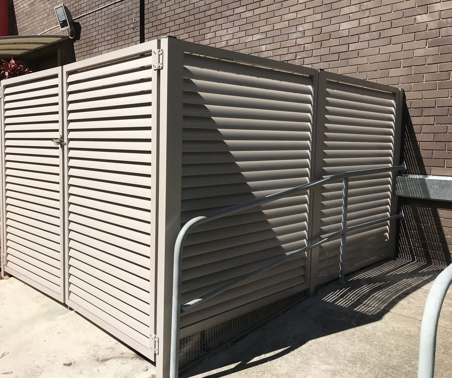 Pale grey aluminium louvre enclosure with double doors screening bins beside a brick building