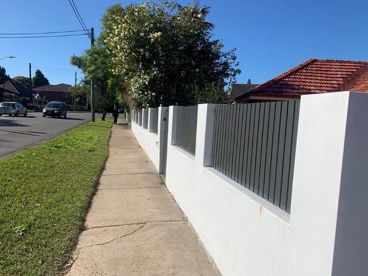 Grey vertical slat infills set into openings in a long white rendered front wall beside a footpath